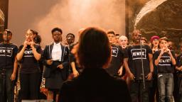 Group of young people singing on stage with conductor in foreground